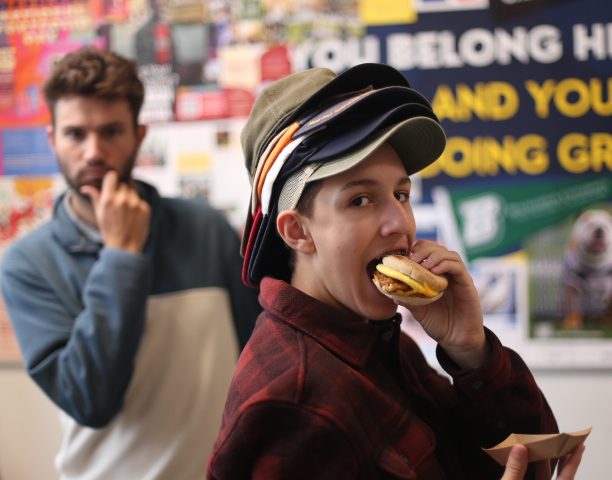 Foster Cardoza stacks multiple hats on his head as he takes a bite out of his breakfast sandwich. Meanwhile, Ryan Silvester secretly poses behind Cardoza. 