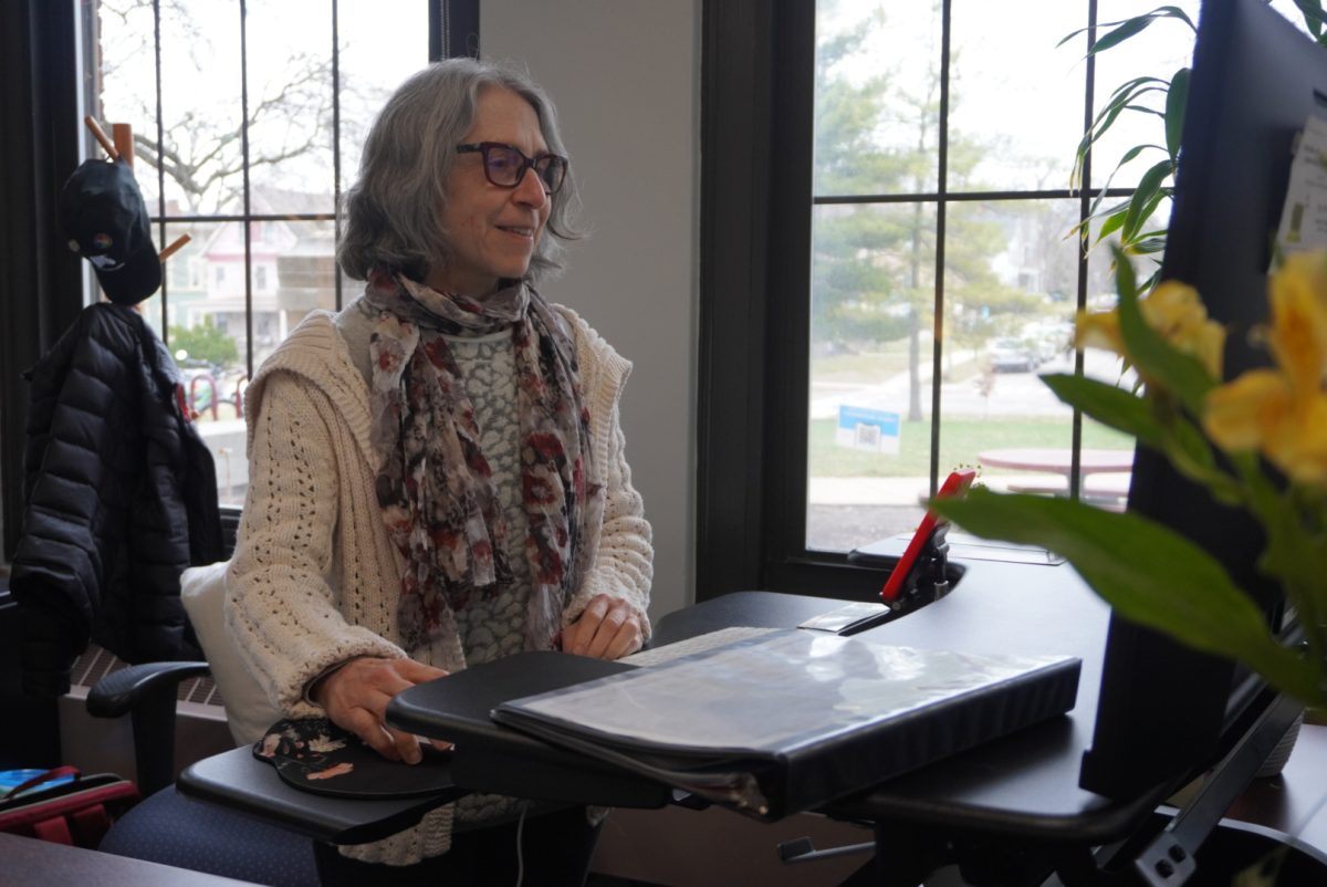 Joann Constantinides stands at her office desk, surrounded by screens, papers and a little greenery.