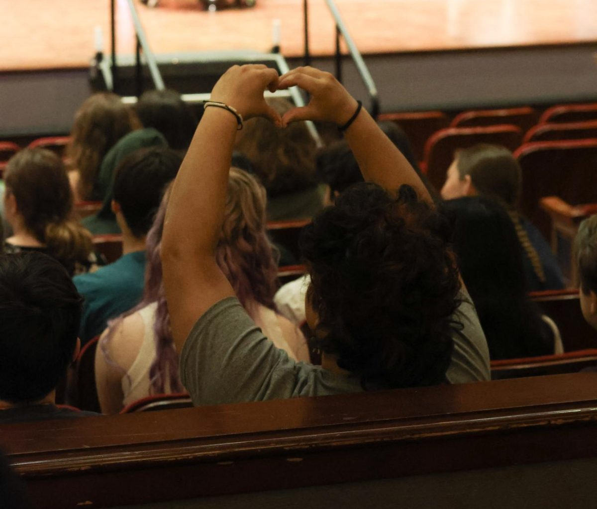 Neila Shee makes a heart with their hands. Shee watches as their friends walk across the stage during the graduation rehearsal.