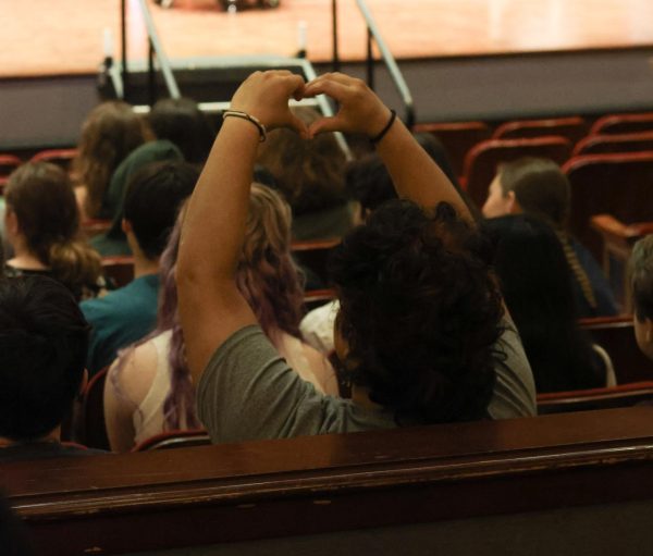 Neila Shee makes a heart with their hands. Shee watches as their friends walk across the stage during the graduation rehearsal.