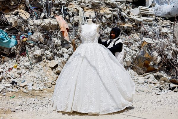 A Palestinian woman looks at a wedding dress displayed outside a shop next to a pile of rubble, as brides in Gaza turn to reused gowns amid soaring prices and shortages caused by the war, in Khan Younis in the southern Gaza Strip, May 9, 2026. REUTERS/Mahmoud Issa