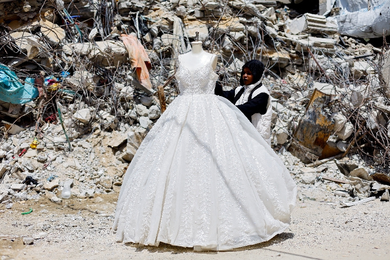 A Palestinian woman looks at a wedding dress displayed outside a shop next to a pile of rubble, as brides in Gaza turn to reused gowns amid soaring prices and shortages caused by the war, in Khan Younis in the southern Gaza Strip, May 9, 2026. REUTERS/Mahmoud Issa